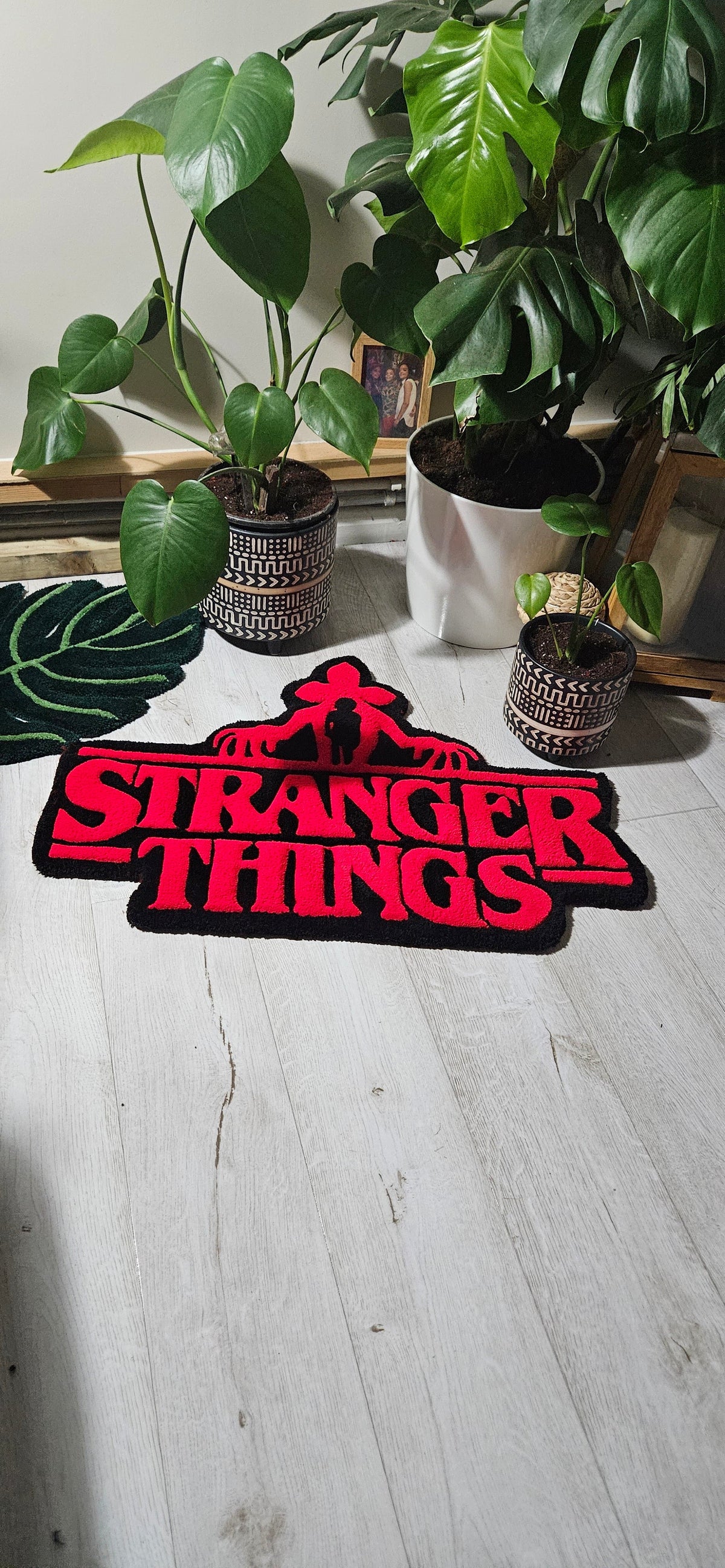 A Stranger Things themed rug featuring the red title logo and a black silhouette of the character Eleven, displayed on a light wood floor next to potted green plants.